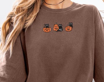 May include: Brown sweatshirt featuring a Halloween design. The design includes three orange pumpkins with black cat illustrations. The cats have various poses, adding a playful touch to the fall-themed apparel.