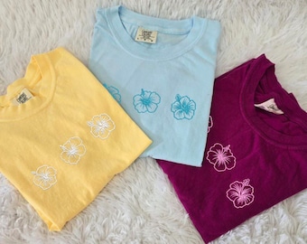 May include: Three t-shirts in yellow, light blue, and purple, each featuring a floral design. The shirts are folded and displayed on a white surface. The floral design is embroidered on the front of each shirt.