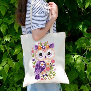 Peut inclure: Un sac fourre-tout en toile blanche avec un hibou violet brod&eacute; portant une couronne florale. Le hibou a de grands yeux bruns et est entour&eacute; de fleurs roses, orange et vertes.