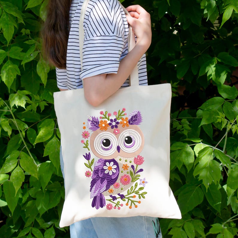 Peut inclure: Un sac fourre-tout en toile blanche avec un hibou violet brod&eacute; portant une couronne florale. Le hibou a de grands yeux bruns et est entour&eacute; de fleurs roses, orange et vertes.