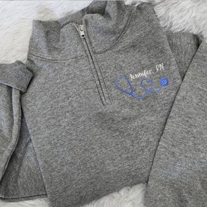 May include: Gray fleece pullover with a zipper and a stethoscope design with hearts. The text "Jennifer, RN" is embroidered on the front.