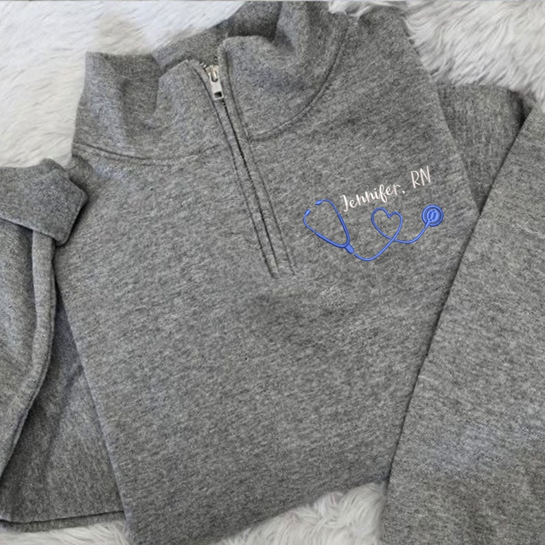 May include: Gray fleece pullover with a zipper and a stethoscope design with hearts. The text "Jennifer, RN" is embroidered on the front.