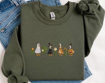 May include: An olive green sweatshirt featuring an embroidered design of four geese dressed in Halloween costumes. The geese are wearing a ghost costume, a witch costume, an orange dress, and a pumpkin costume. The design includes flowers.