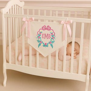 Puede incluir: Una cuna blanca con una pancarta crema bordada con las iniciales "EMB" dentro de una corona floral. La pancarta está adornada con lazos rosas. Un bebé duerme plácidamente dentro de la cuna, que tiene una sábana con estampado floral.