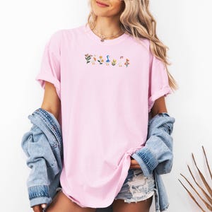 May include: A pink oversized t-shirt with an embroidered design of flowers, ducks, and mushrooms.