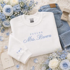 May include: A white crewneck sweatshirt featuring light blue embroidery that reads FUTURE Mrs. Brown on the chest and the initials T and H separated by a heart on the sleeve cuff. The garment is displayed alongside blue jeans and a ring box.