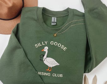 May include: An olive green sweatshirt with the words "SILLY GOOSE NURSING CLUB" and a cartoon goose wearing a nurse's cap and stethoscope. A gold necklace and a brown hat are also visible.