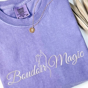 May include: A lavender colored t-shirt with the text "Boudoir Magic" in white cursive lettering. The shirt has a tag that reads "Comfort Colors".