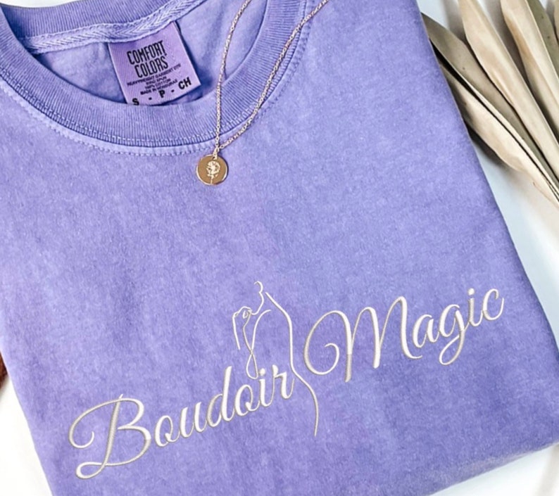 May include: A lavender colored t-shirt with the text "Boudoir Magic" in white cursive lettering. The shirt has a tag that reads "Comfort Colors".