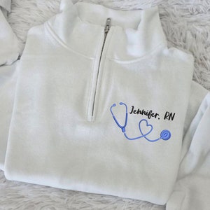May include: White quarter-zip sweatshirt with a blue stethoscope and heart graphic. The text "Jennifer. RN" is printed on the sweatshirt.