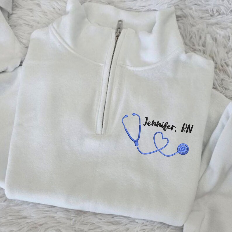 May include: White quarter-zip sweatshirt with a blue stethoscope and heart graphic. The text "Jennifer. RN" is printed on the sweatshirt.