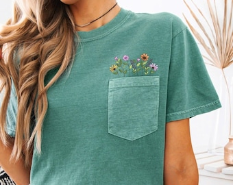 May include: A teal green t-shirt with a pocket featuring an embroidered design of colorful flowers.
