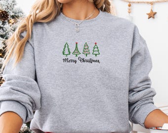 May include: A light gray sweatshirt with a festive Christmas design. The design features four green Christmas trees with a red star on one, and the words "Merry Christmas," in black script. The sweatshirt is a comfortable, casual style.