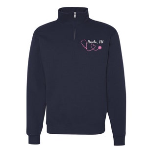 May include: Navy blue quarter-zip sweatshirt with a pink stethoscope and heart graphic and the text "Nicole, RN".