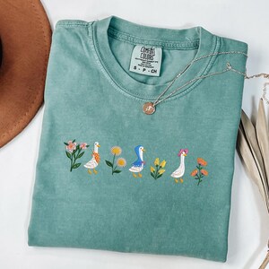 May include: A teal blue t-shirt with a white embroidered design of three ducks and flowers. The shirt has a tag that says "Comfort Colors" and "S - P - CH".