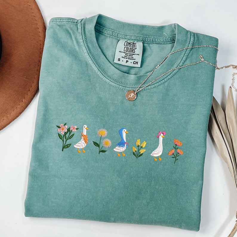 May include: A teal blue t-shirt with a white embroidered design of three ducks and flowers. The shirt has a tag that says "Comfort Colors" and "S - P - CH".