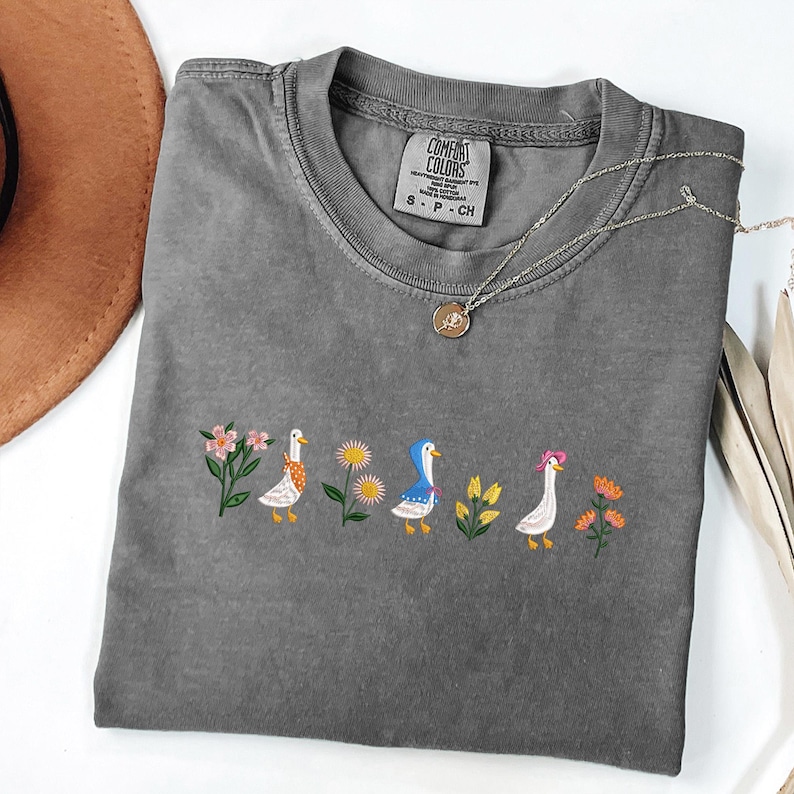 May include: A gray t-shirt with embroidered ducks and flowers. The shirt has a Comfort Colors tag with the text "Comfort Colors" and "S - P - CH".