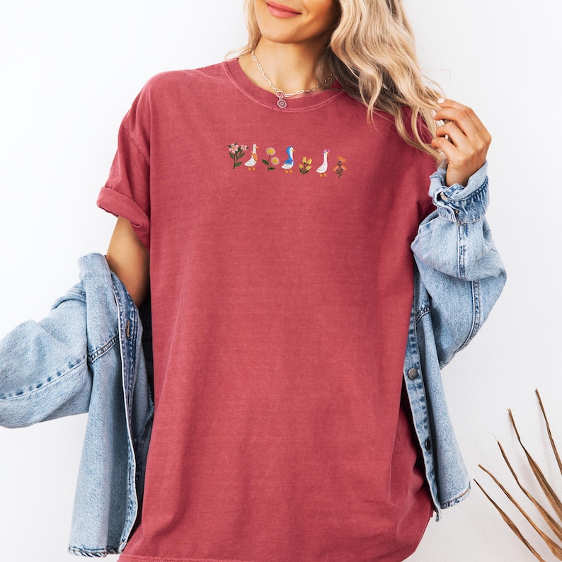 May include: A maroon t-shirt with a small embroidered design of flowers and ducks. The design is in shades of blue, yellow, and green.