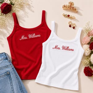 May include: A red tank top and a white tank top, both featuring the embroidered text Mrs. Williams in a cursive script. The red top has white lettering, while the white top has red lettering. Both garments have wide shoulder straps and a scoop neckline.