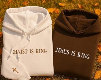 May include: A white hooded sweatshirt with the brown text CHRIST IS KING and a small brown cross on the sleeve, alongside a dark brown hooded sweatshirt with the white text JESUS IS KING, both featuring matching drawstrings.