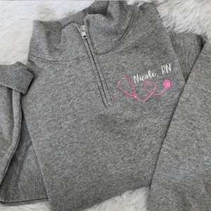 May include: Gray fleece pullover with a zipper and a pink stethoscope graphic with the text "Nicole, RN".