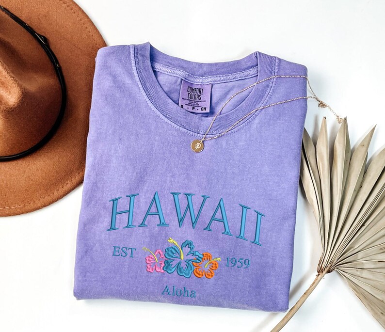 May include: A purple t-shirt with the text "HAWAII EST 1959 Aloha" and a floral design. The shirt is made by Comfort Colors.