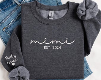 May include: A dark gray sweatshirt with the text "mimi est. 2024" in white lettering. The sweatshirt is folded and displayed on a white surface with a pair of blue jeans.