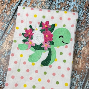 May include: A green turtle with a pink, white, and green flower bouquet on its back. The turtle is embroidered on a white fabric with pink, yellow, and green polka dots.