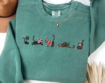 May include: Teal green sweatshirt with embroidered black cats in festive holiday attire. The cats are decorated with Santa hats, Christmas lights, and a red and white outfit. A gold necklace and a brown hat are also visible.