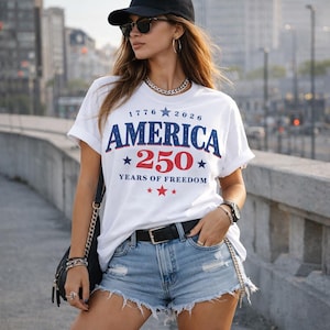 Puede incluir: Una camiseta blanca con el texto "AMERICA 250 YEARS OF FREEDOM" en azul y rojo. La camiseta tiene cuello redondo y mangas cortas. La persona lleva pantalones cortos vaqueros, un cinturón negro y una gorra de béisbol negra.