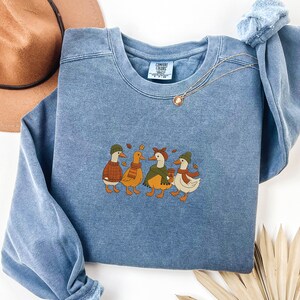 May include: A blue Comfort Colors sweatshirt featuring an embroidered design of four ducks in fall attire. The ducks are wearing hats, scarves, and sweaters, and one is holding a coffee cup. A gold necklace and a brown hat are also visible.