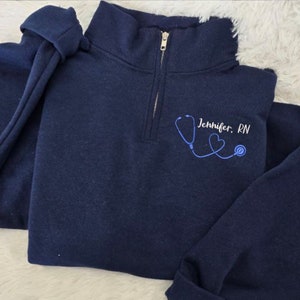 May include: A navy blue quarter-zip sweatshirt with a blue stethoscope and two hearts embroidered on the front. The text "Jennifer. RN" is also embroidered on the sweatshirt.