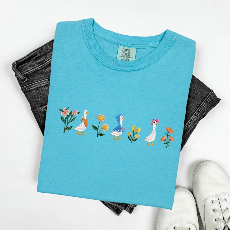 May include: A light blue t-shirt with an embroidered design of four geese walking in a line. The geese are wearing different colored hats and are surrounded by flowers. The t-shirt has a small tag that says "Comfort Colors" on it.