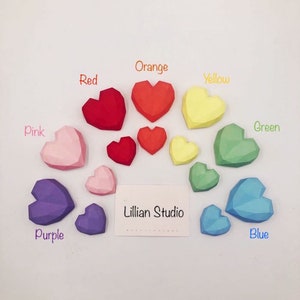 May include: A set of 10 colourful paper hearts in various sizes, each with a geometric design. The hearts are arranged in a rainbow pattern, with the colours pink, purple, blue, green, yellow, orange, and red. The hearts are labelled with their corresponding colours. The text "Lillian Studio" is printed on a white card in the centre of the image.