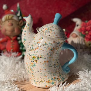 May include: A white ceramic teapot with a blue lid and handle. The teapot is decorated with a splatter paint design in orange, yellow, green, and black.