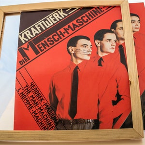 May include: A framed album cover featuring the band Kraftwerk. The album cover is predominantly red with the band's name and album title in white and black text. The cover art depicts four men in red shirts and black ties. The frame is made of light-colored wood.