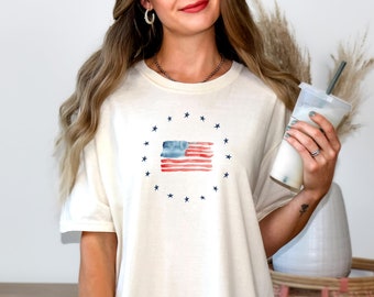 May include: A white oversized t-shirt with a watercolor American flag design in the center. The flag is surrounded by a circle of blue stars.