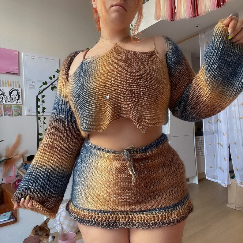 May include: A two-piece knitted outfit in shades of blue and brown. The top is a cropped sweater with long sleeves and shoulder straps. The matching skirt has a drawstring waist and a crocheted hem. The outfit is handmade.