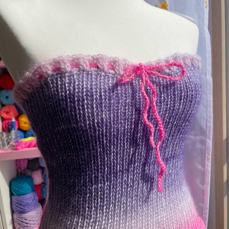 May include: A knitted tube top in shades of purple and pink, with a gradient effect. The top features a pink, ruffled trim at the top and a pink bow detail. The garment is displayed on a white mannequin.