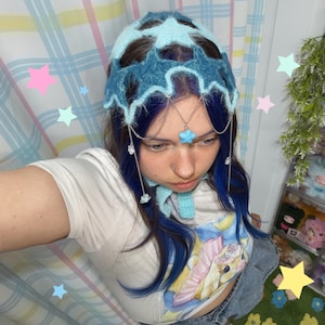 May include: A fuzzy, light blue and dark blue star-patterned crochet headpiece featuring silver-toned chains that drape across the forehead. A single, small, light blue star charm hangs from the center of the chain, with additional small star charms dangling from the sides of the headpiece.