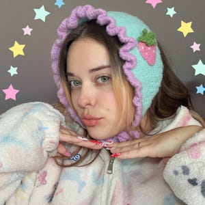 May include: A light blue crochet bonnet with a ruffled lavender trim and a pink strawberry applique. The bonnet is handmade and features a soft, textured appearance. The bonnet is a unique and stylish accessory.