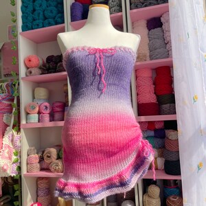 May include: A strapless, knitted dress in a gradient of colors, from purple to pink. The dress has a ruffled top edge and a drawstring detail on the side. The dress is displayed on a mannequin, with shelves of yarn in the background.