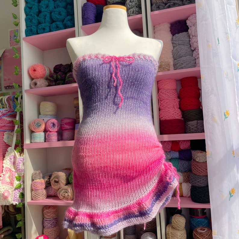 May include: A strapless, knitted dress in a gradient of colors, from purple to pink. The dress has a ruffled top edge and a drawstring detail on the side. The dress is displayed on a mannequin, with shelves of yarn in the background.