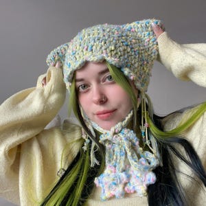 以下が含まれることがあります： パステルイエロー、ブルー、ピンクの手作りの猫耳かぎ針編み帽子。帽子には、フリルの縁とビーズのアクセントが付いたお揃いのスカーフが付いています。帽子はあごの下で結ばれています。