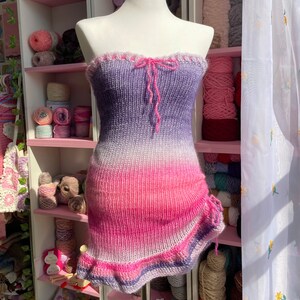 May include: A strapless, knitted dress in a gradient of purple, white, and pink. The top edge has a ruffled trim and a pink bow. The dress has a fitted silhouette with a ruffled asymmetrical hemline. The dress is displayed on a mannequin.