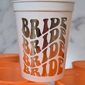 A white plastic cup with the word "BRIDE" in a retro font, in shades of gold and orange. The cup is surrounded by other orange cups. The design is repeated multiple times down the side of the cup. Great for bachelorette parties.