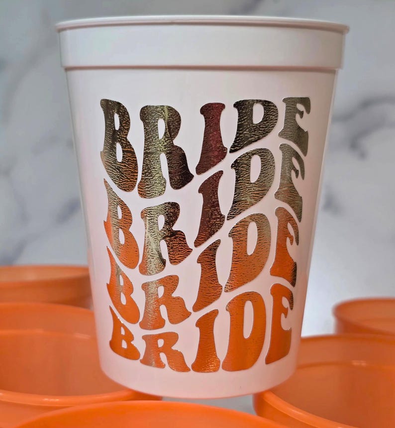 A white plastic cup with the word "BRIDE" in a retro font, in shades of gold and orange. The cup is surrounded by other orange cups. The design is repeated multiple times down the side of the cup. Great for bachelorette parties.