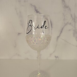 Może przedstawiać: Przezroczysty kieliszek do wina ze słowem "Bride" w czarnym skrypcie. Dolna połowa kieliszka pokryta jest białym brokatem. N&oacute;żka i podstawa kieliszka są r&oacute;wnież pokryte białym brokatem.