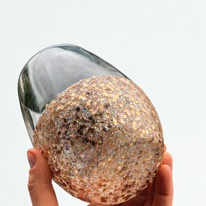 A clear glass wine glass with a glitter-covered base. The glitter is a mix of gold and iridescent colors, creating a sparkling effect. The glass is held against a white background.