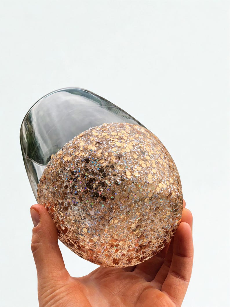 A clear glass wine glass with a glitter-covered base. The glitter is a mix of gold and iridescent colors, creating a sparkling effect. The glass is held against a white background.